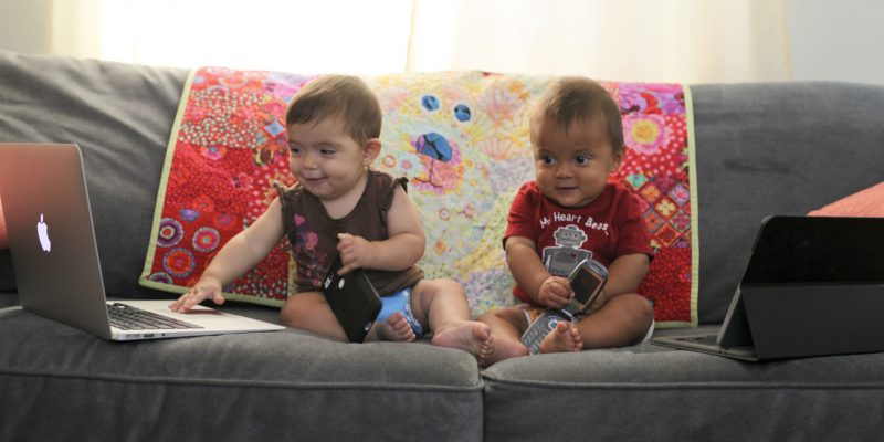 Two toddlers sitting on a couch with cell phones and computers.