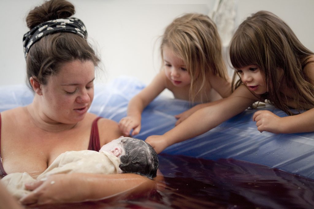 Two big sisters are reaching to touch their new baby that was just born in a birth pool. Mom holds baby.