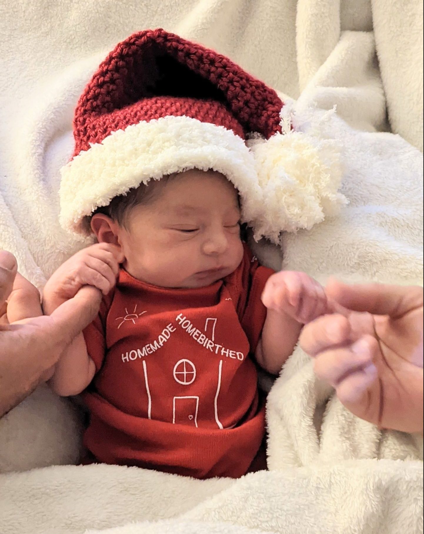 Newborn wearing a crochet santa hat, a onesie that says "Homemade Homebirthed". and holding onto two adult fingers.